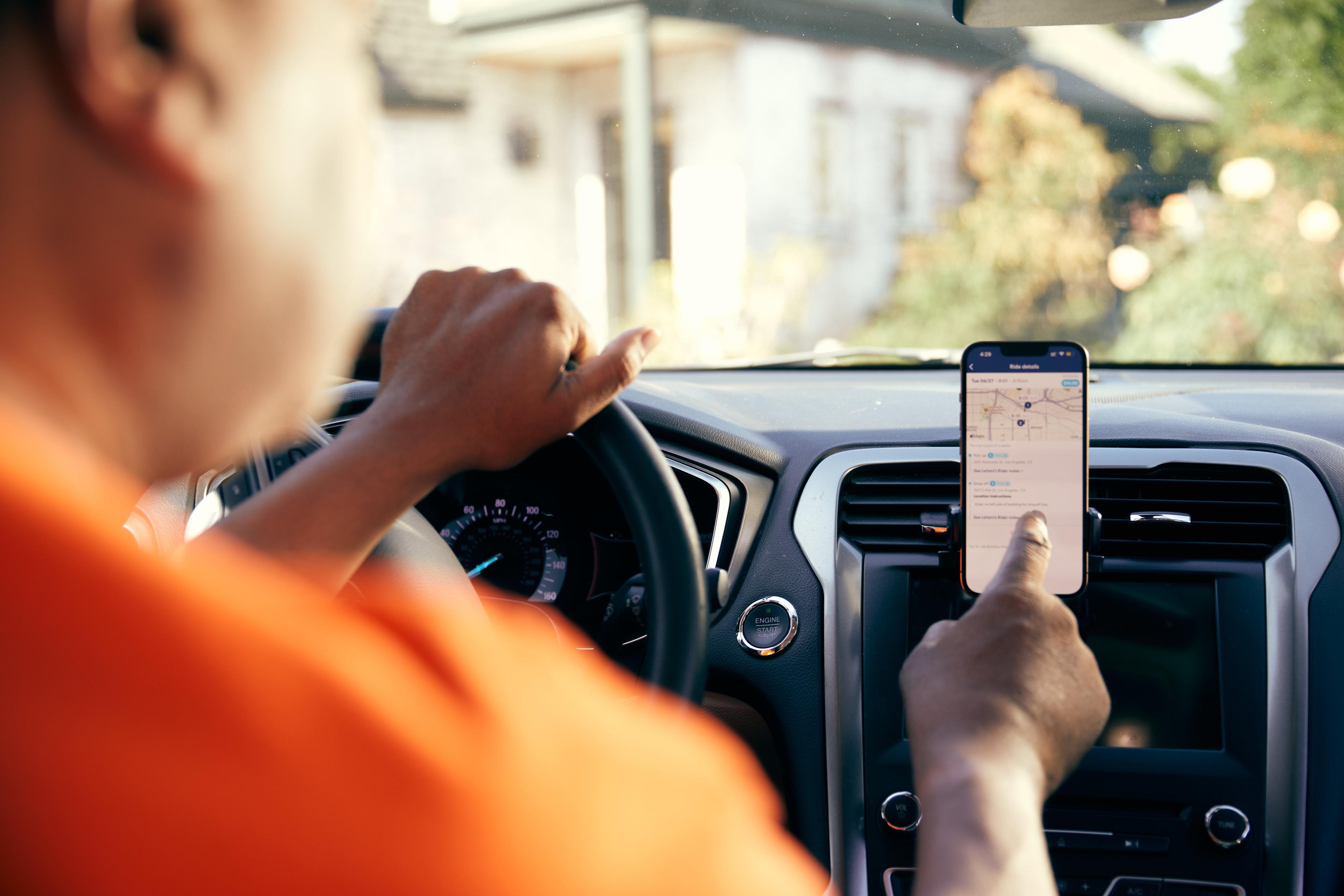 Driver in orange shirt touching smartphone mounted on car dashboard displaying navigation map interface