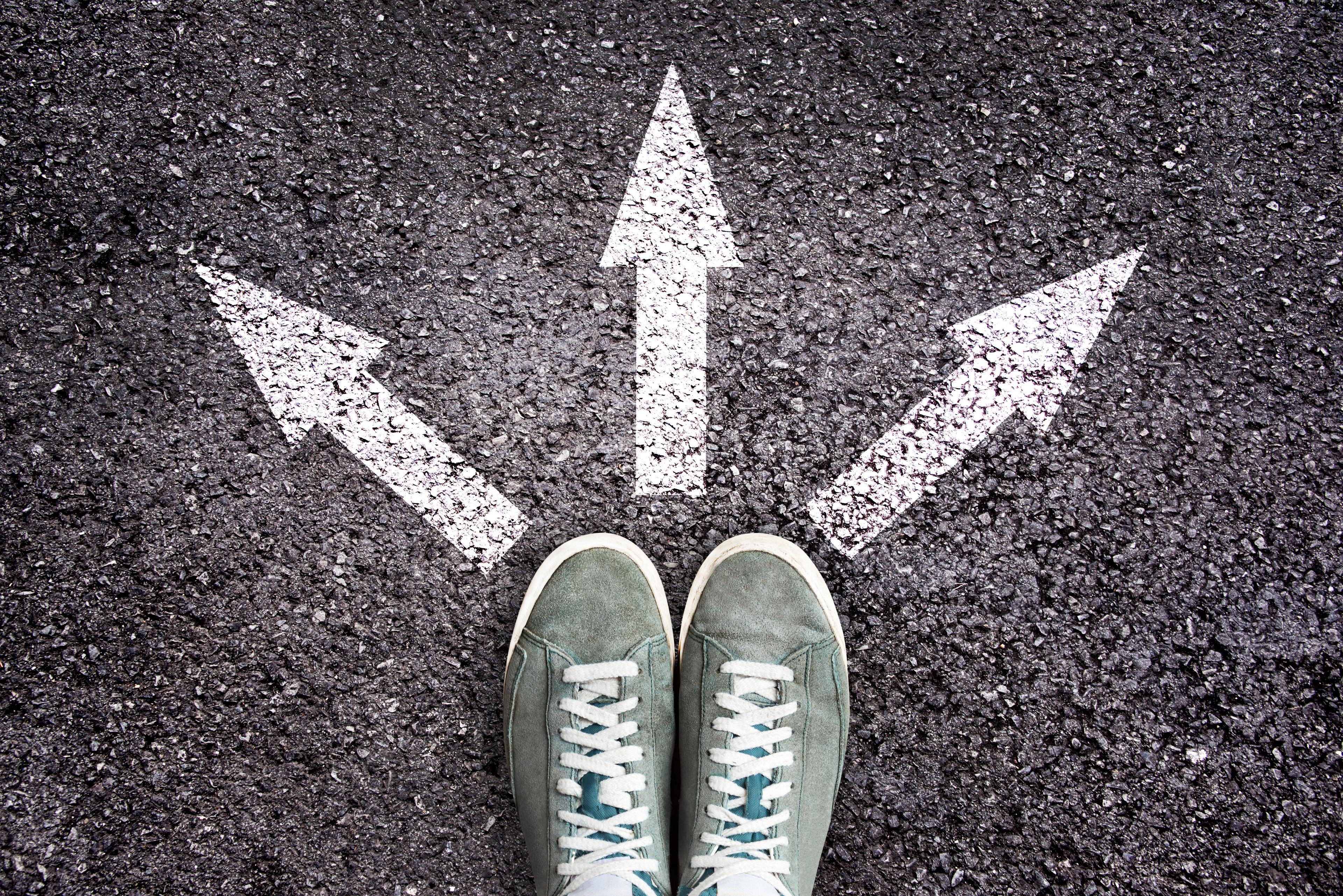 Gray sneakers standing at intersection of three white painted arrows pointing left, straight ahead, and right on asphalt surface