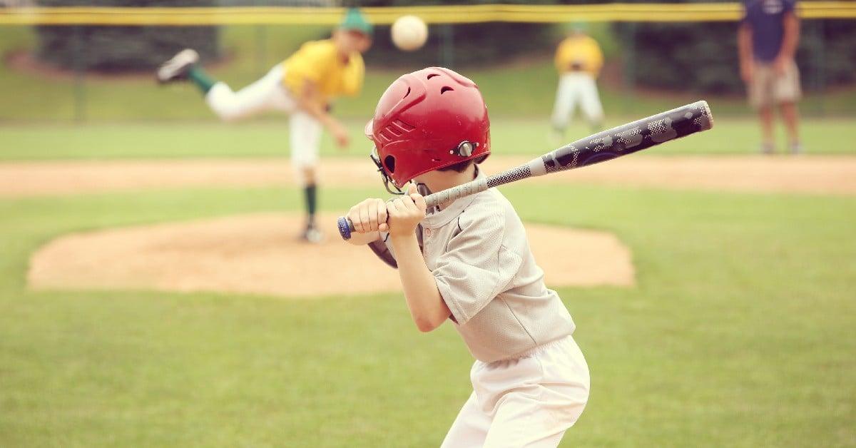 Young baseball player in batting stance wearing red helmet and white uniform, with pitcher visible in background