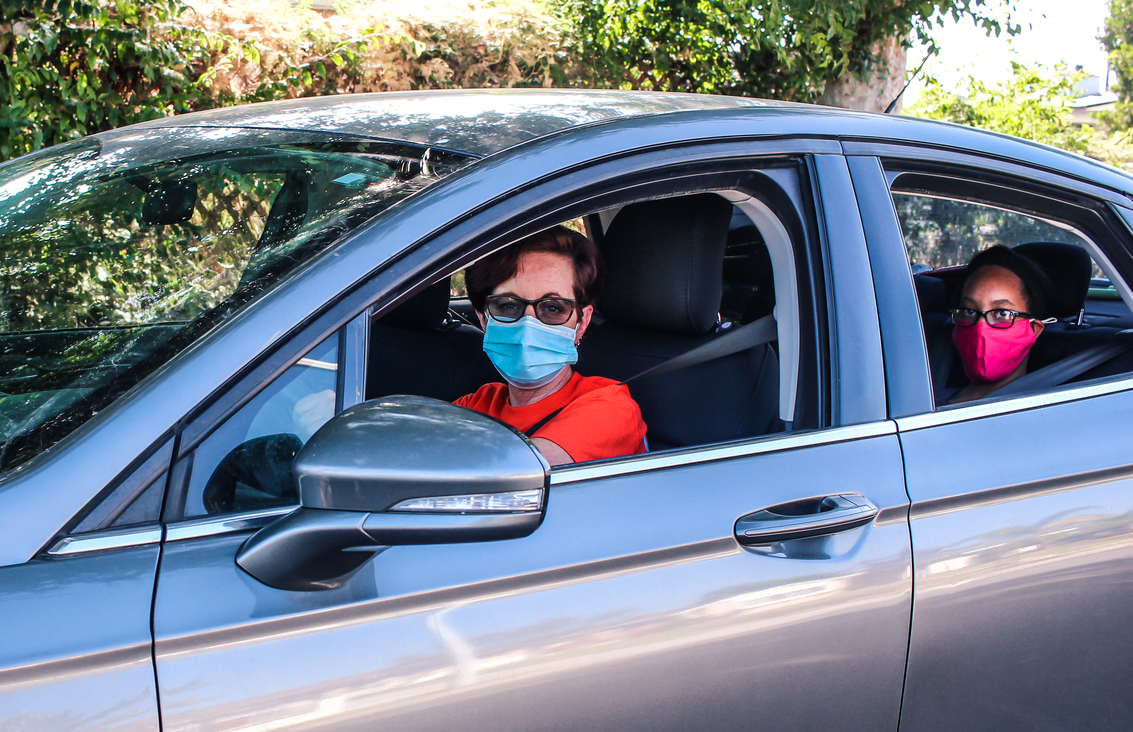 Two people wearing face masks seated in a silver car, driver in orange shirt and passenger in back wearing pink mask