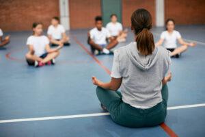 Instructor leading a meditation session with students sitting cross-legged on a blue sports court floor