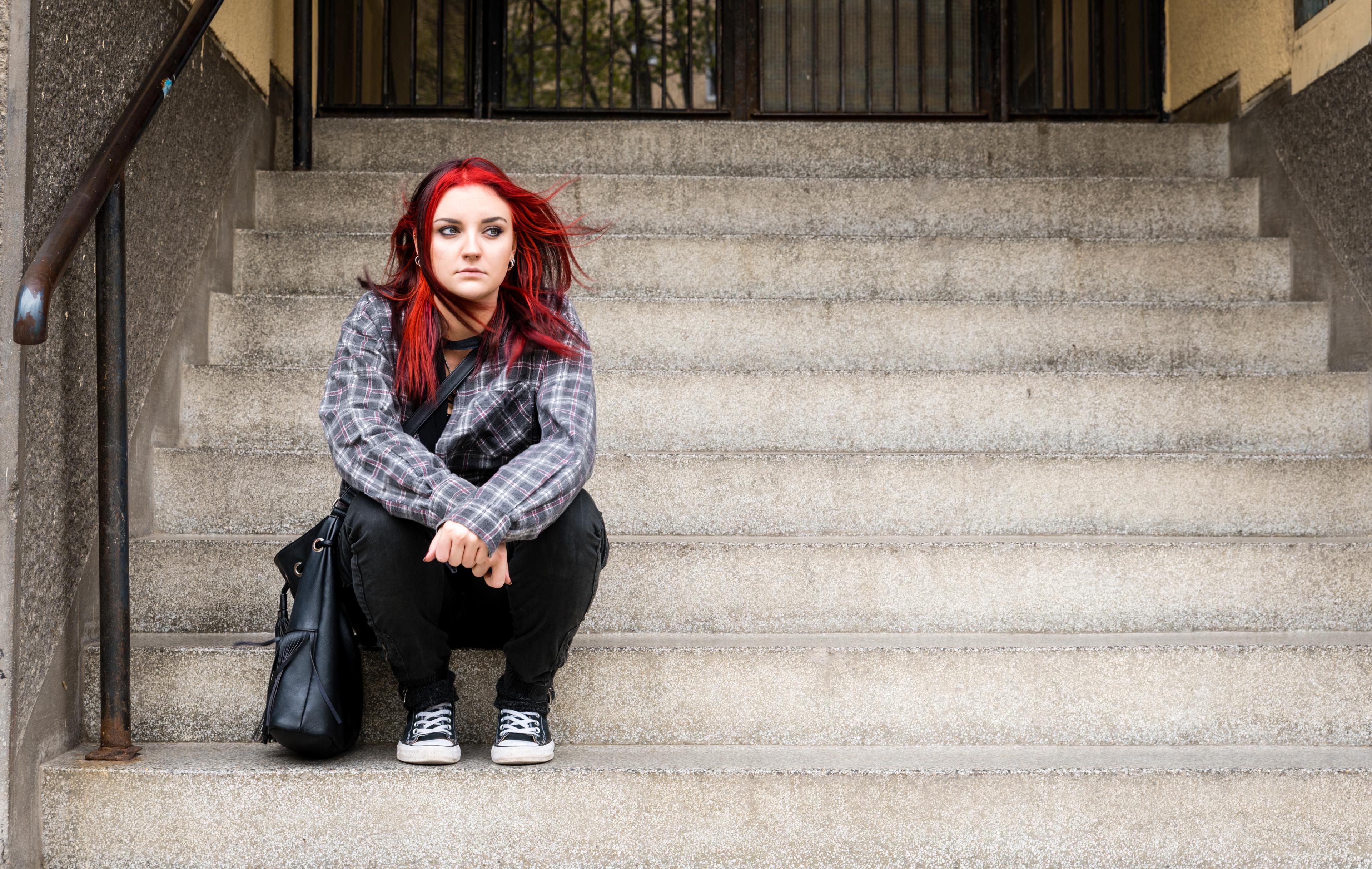 Young person with bright red hair wearing plaid shirt and black pants, crouching on concrete steps with urban backdrop