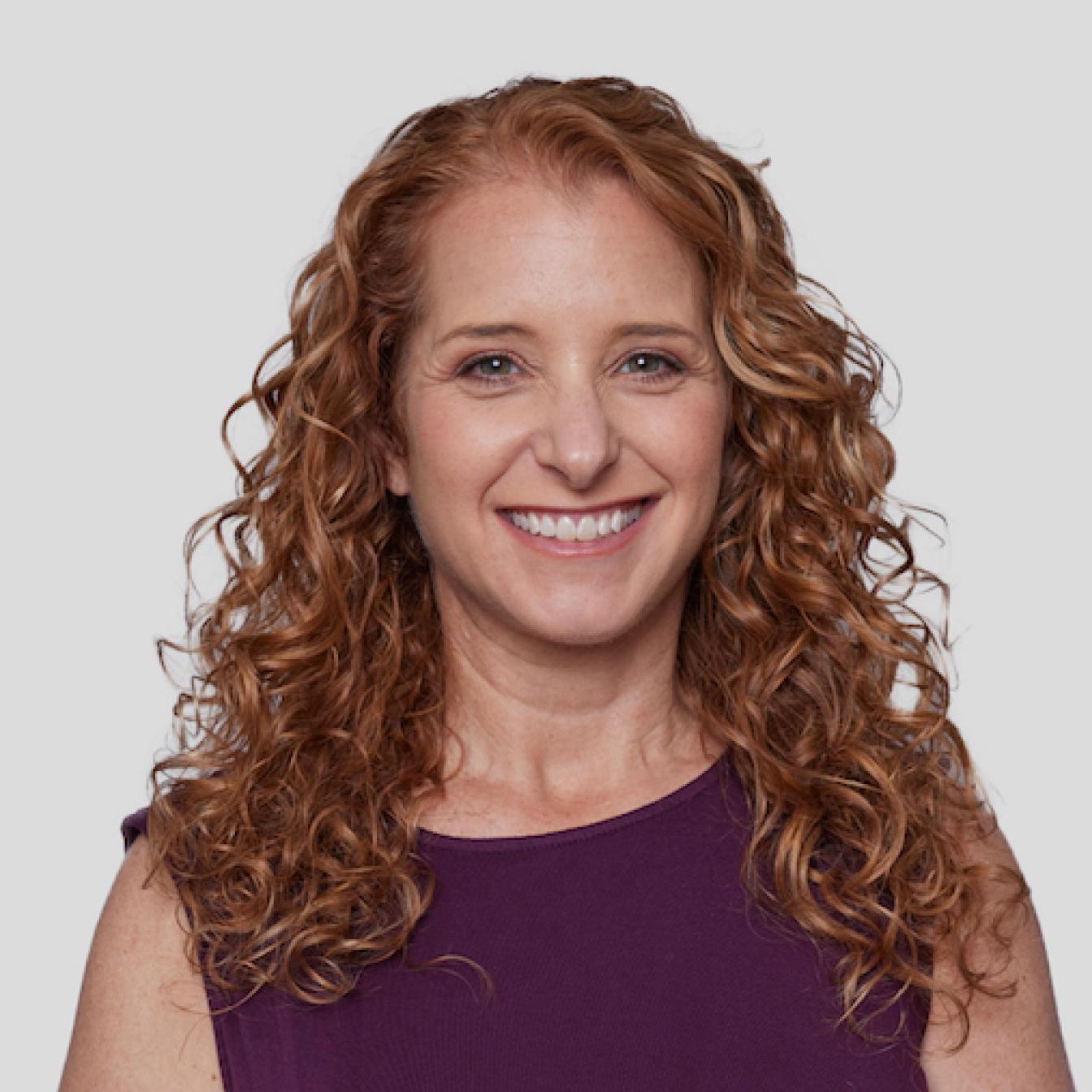 Professional headshot of a person with long curly auburn hair wearing a purple sleeveless top, smiling warmly against a light background