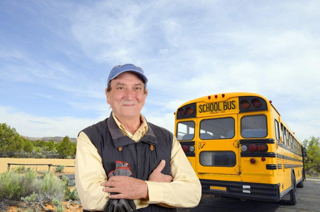 School bus driver wearing cap and vest standing confidently in front of yellow school bus against desert landscape and blue sky