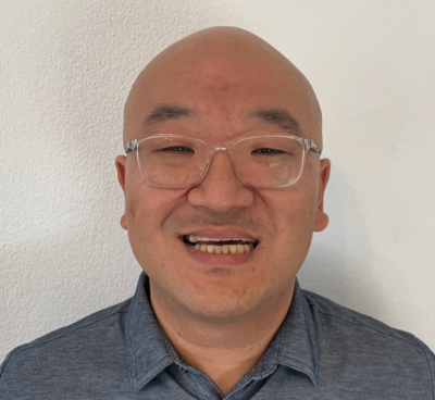 Professional headshot of a person wearing clear-frame glasses and a grey collared shirt, smiling warmly against a white background