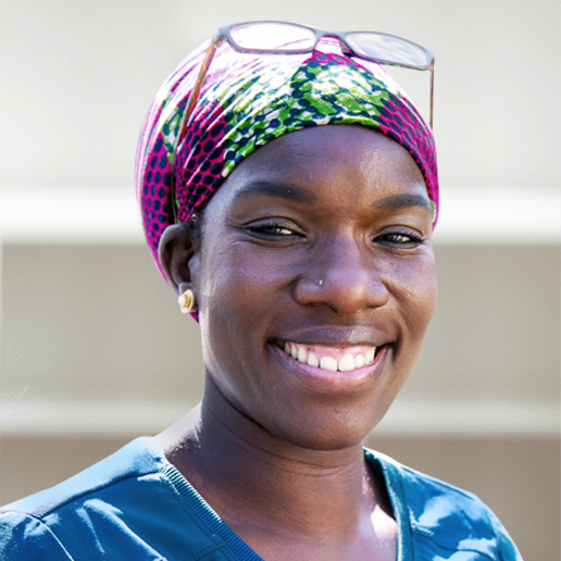 Healthcare worker wearing blue scrubs and vibrant pink-green patterned headwrap, with sunglasses perched on top, showing warm smile