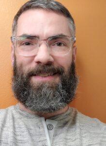 Close-up portrait of a person with a full beard wearing glasses and a grey henley shirt against an orange background