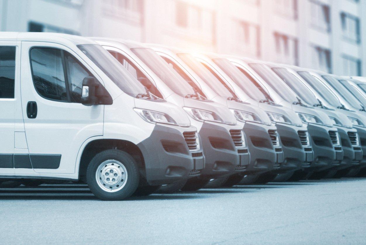 Row of identical white commercial delivery vans parked in line against an urban building background