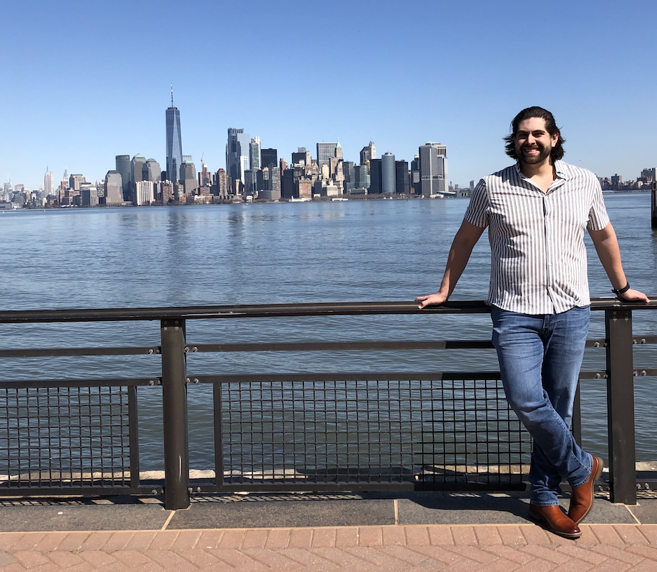 Person standing at waterfront railing with Manhattan skyline view featuring One World Trade Center and downtown buildings across water