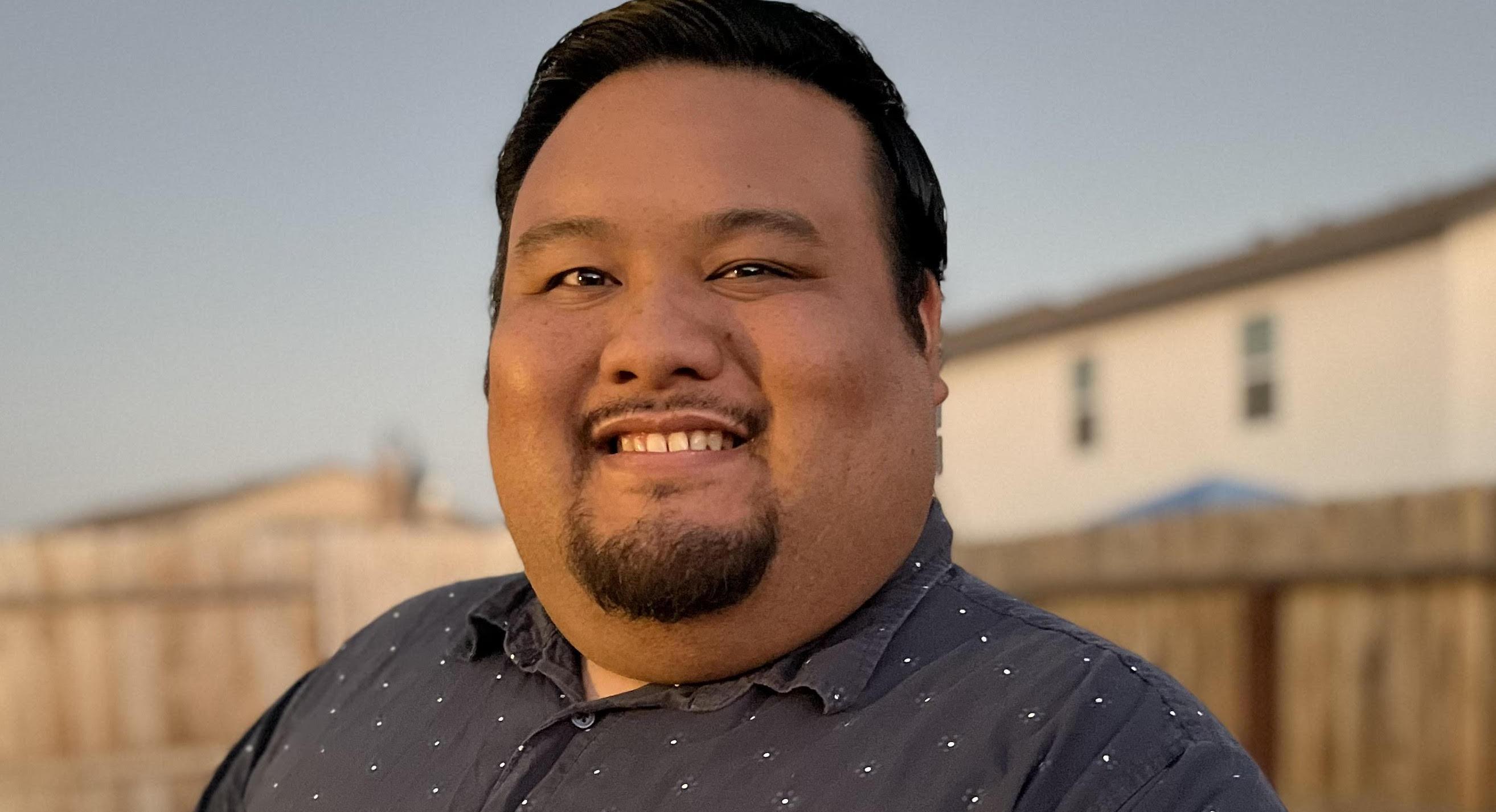 Close-up portrait of a smiling Asian man wearing a dark polka dot shirt, photographed during golden hour with a wooden fence behind