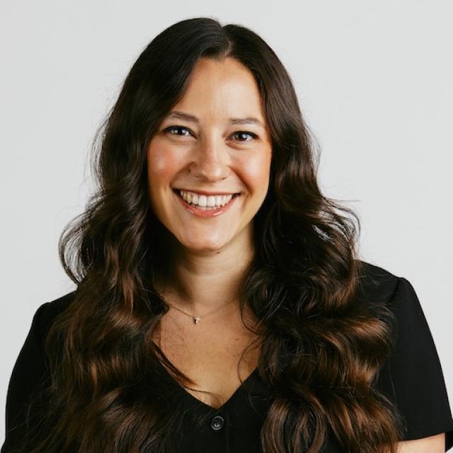 Professional headshot of a woman with long dark wavy hair wearing a black shirt, smiling warmly against a white background