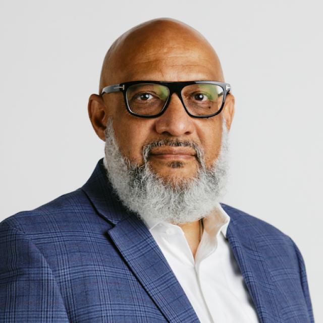 Professional headshot of a person wearing glasses and a blue plaid blazer with a white shirt against a light background