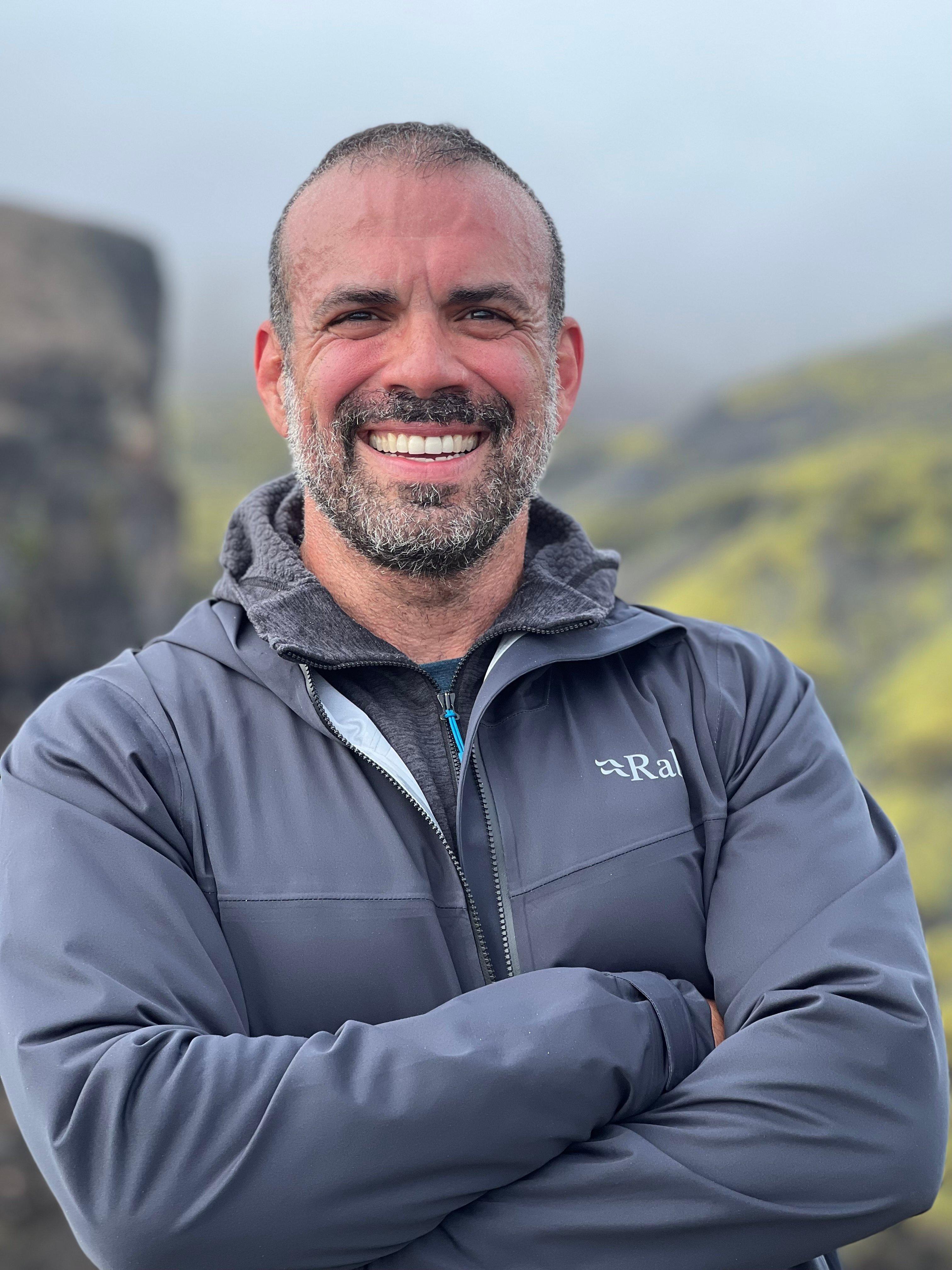 Portrait of man with beard wearing grey RAI jacket outdoors, smiling warmly with crossed arms against mountain backdrop