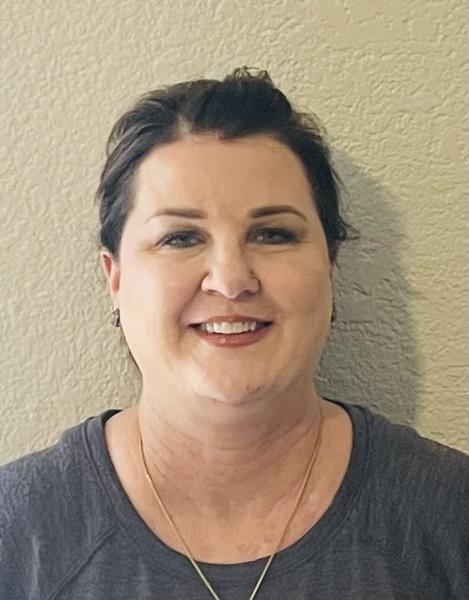 Professional headshot of a person wearing a gray top and necklace, smiling warmly against a textured beige wall