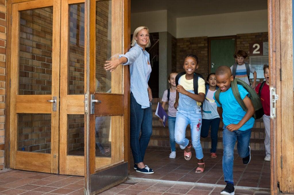 Teacher holding door as excited elementary students exit school building through wooden doorway