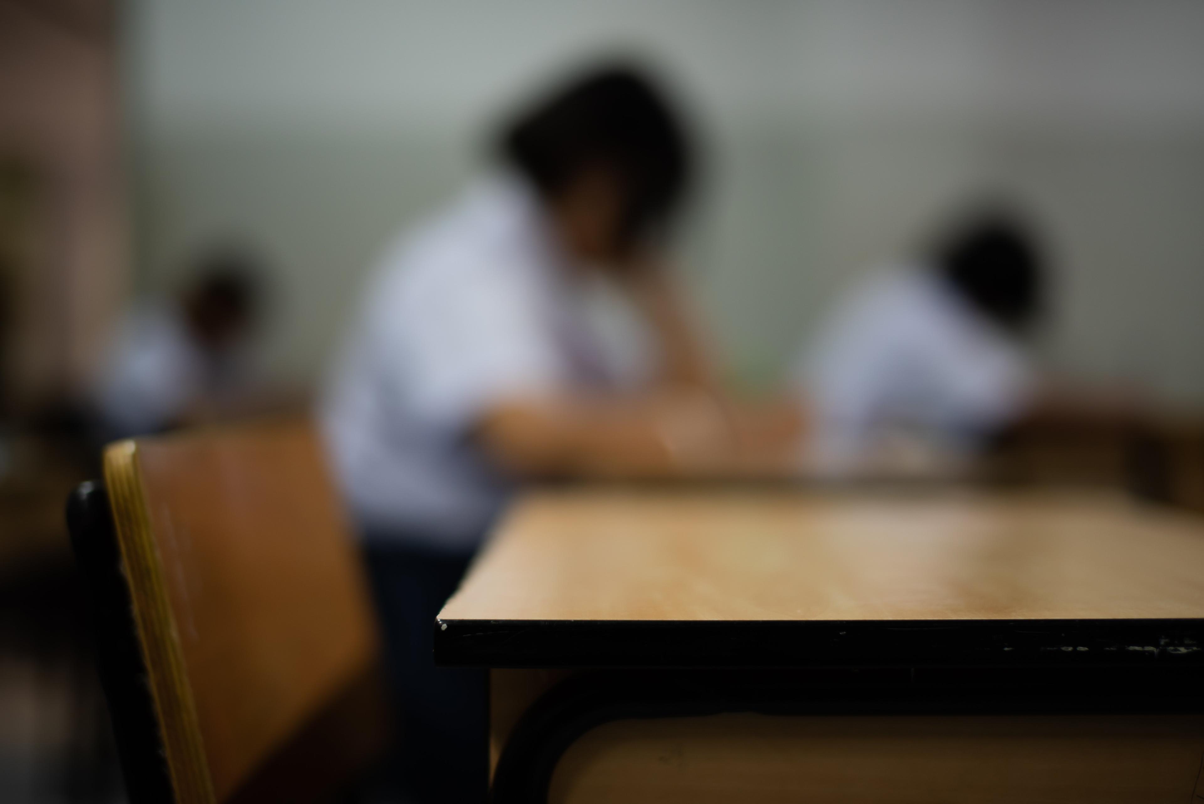 Blurred view of students in white uniforms taking an exam, with desk in foreground