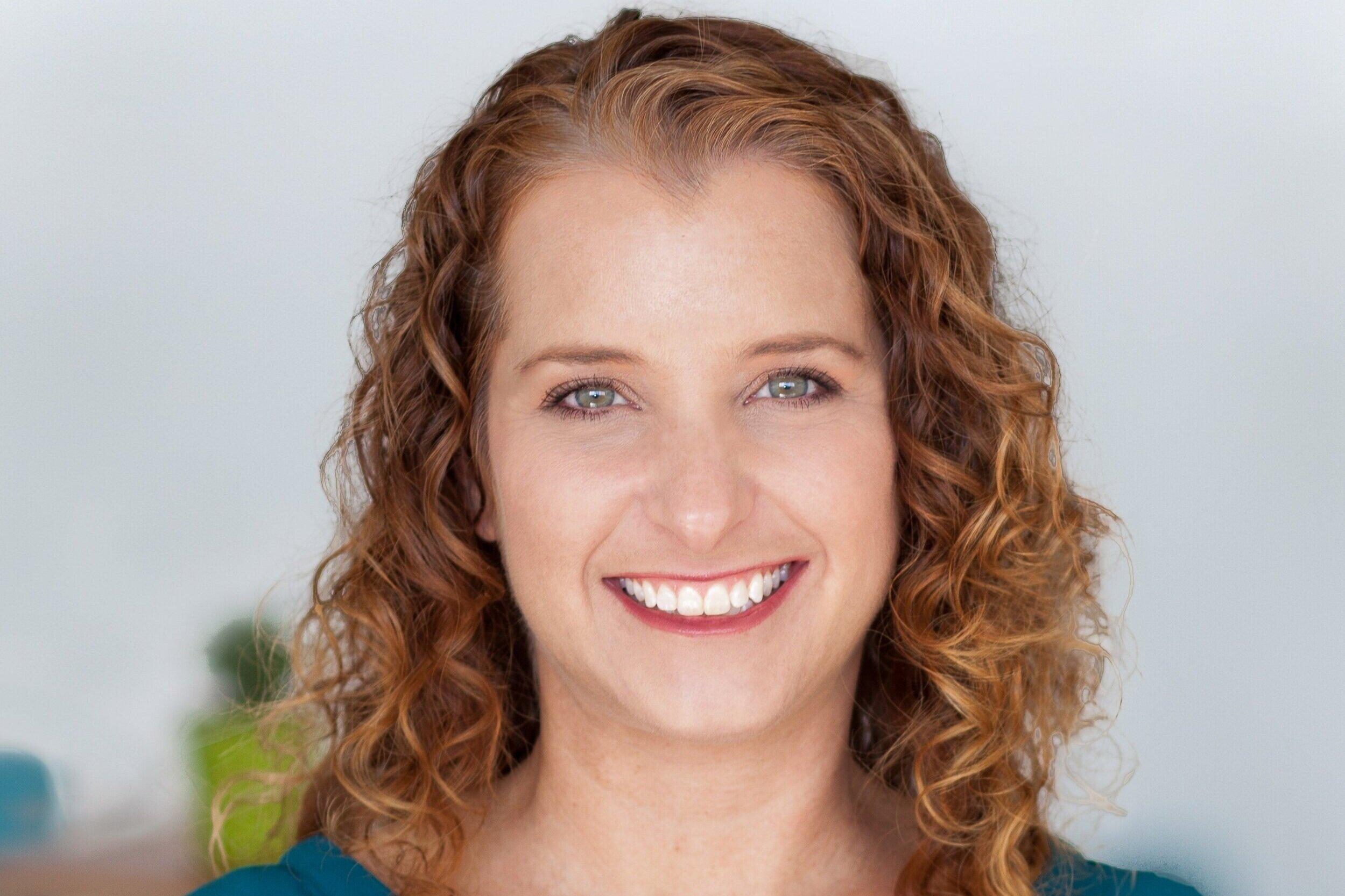 Professional headshot of a person with curly brown hair wearing a teal top, showing a warm genuine smile against a light background