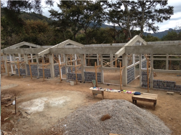 Partially constructed buildings with walls made of recycled plastic bottles, wooden frames, and white roofing in a mountainous setting