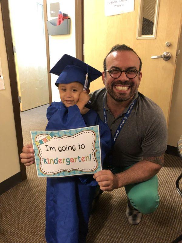 Young graduate in blue cap and gown holds 'I'm going to kindergarten!' sign while posing with educator in school hallway