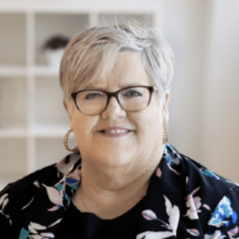 Professional portrait of a person with short silver hair, glasses, and a black floral blouse, smiling warmly against a light background