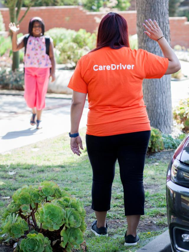 A care provider wearing an orange CareDriver t-shirt waves goodbye to a client on a sunny residential pathway