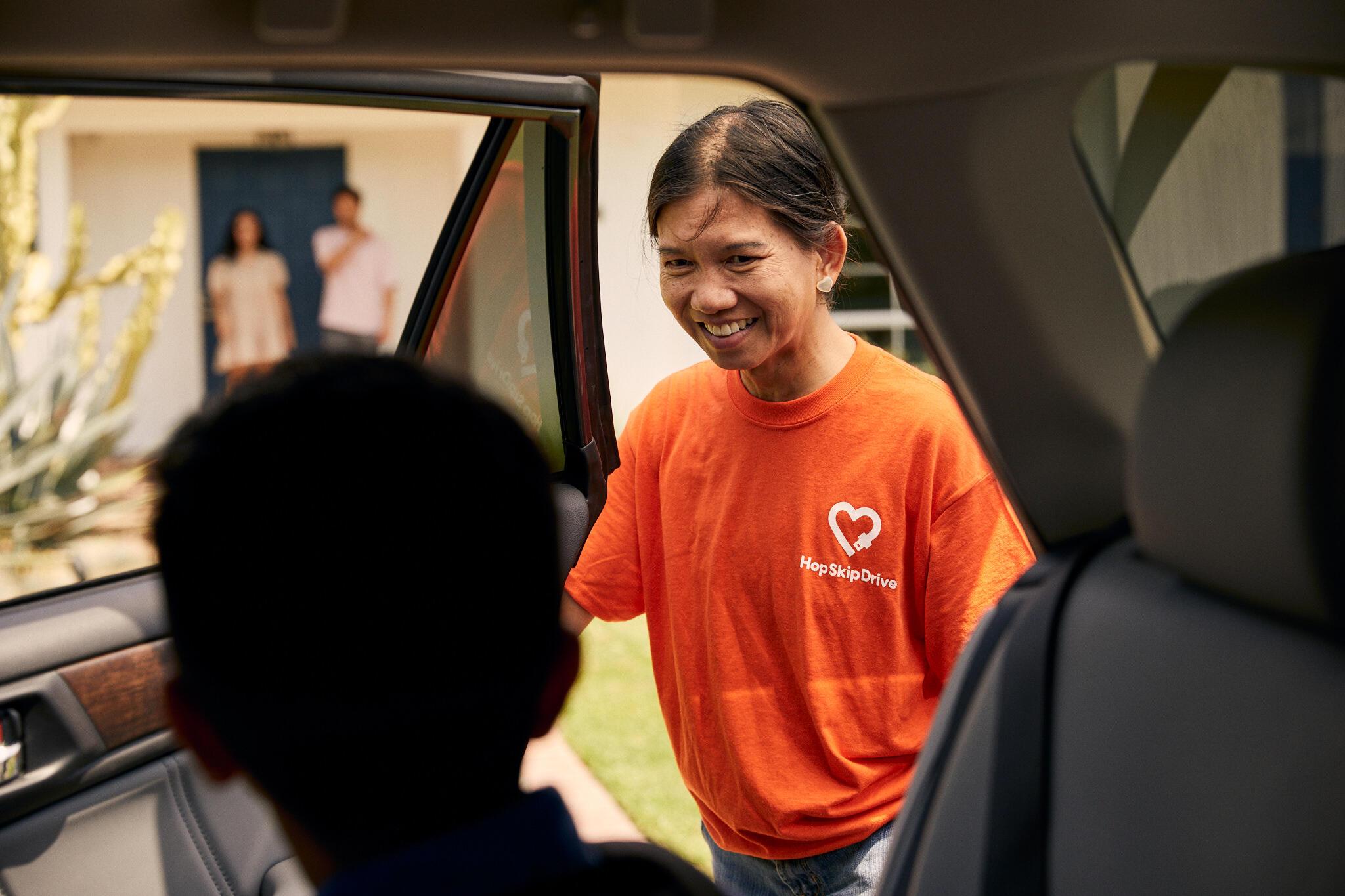 A smiling driver in an orange HopSkipDrive t-shirt stands at a car door, greeting a passenger