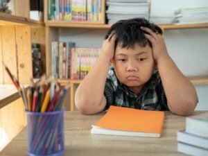Young student with hands on head showing frustration while sitting at desk with books and school supplies