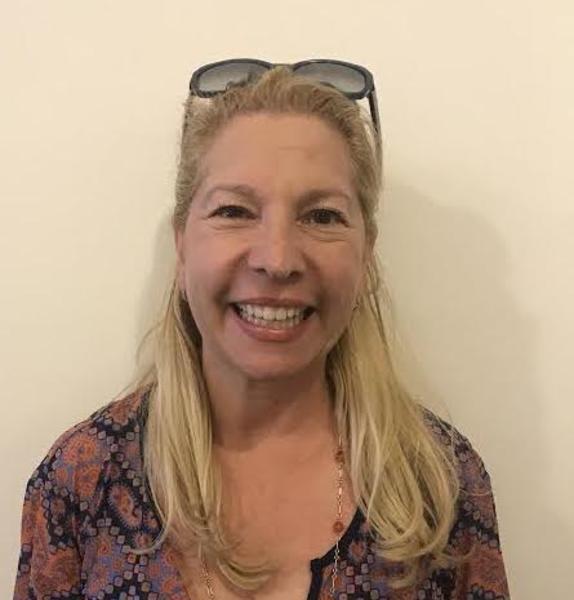Professional headshot of a woman with blonde hair wearing a patterned blouse and sunglasses on head, showing a warm, genuine smile
