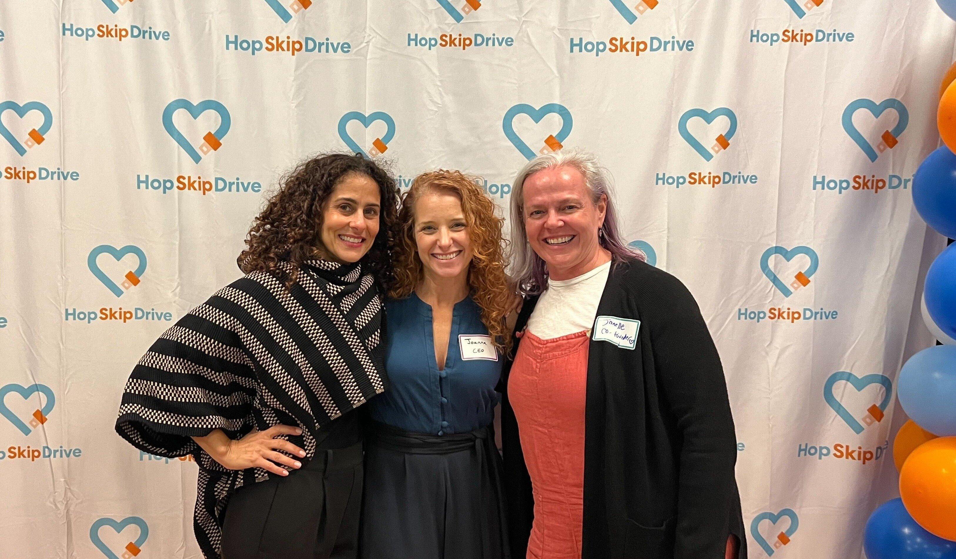 Three women posing in front of HopSkipDrive branded backdrop with logo and heart symbols, with blue and orange balloons visible