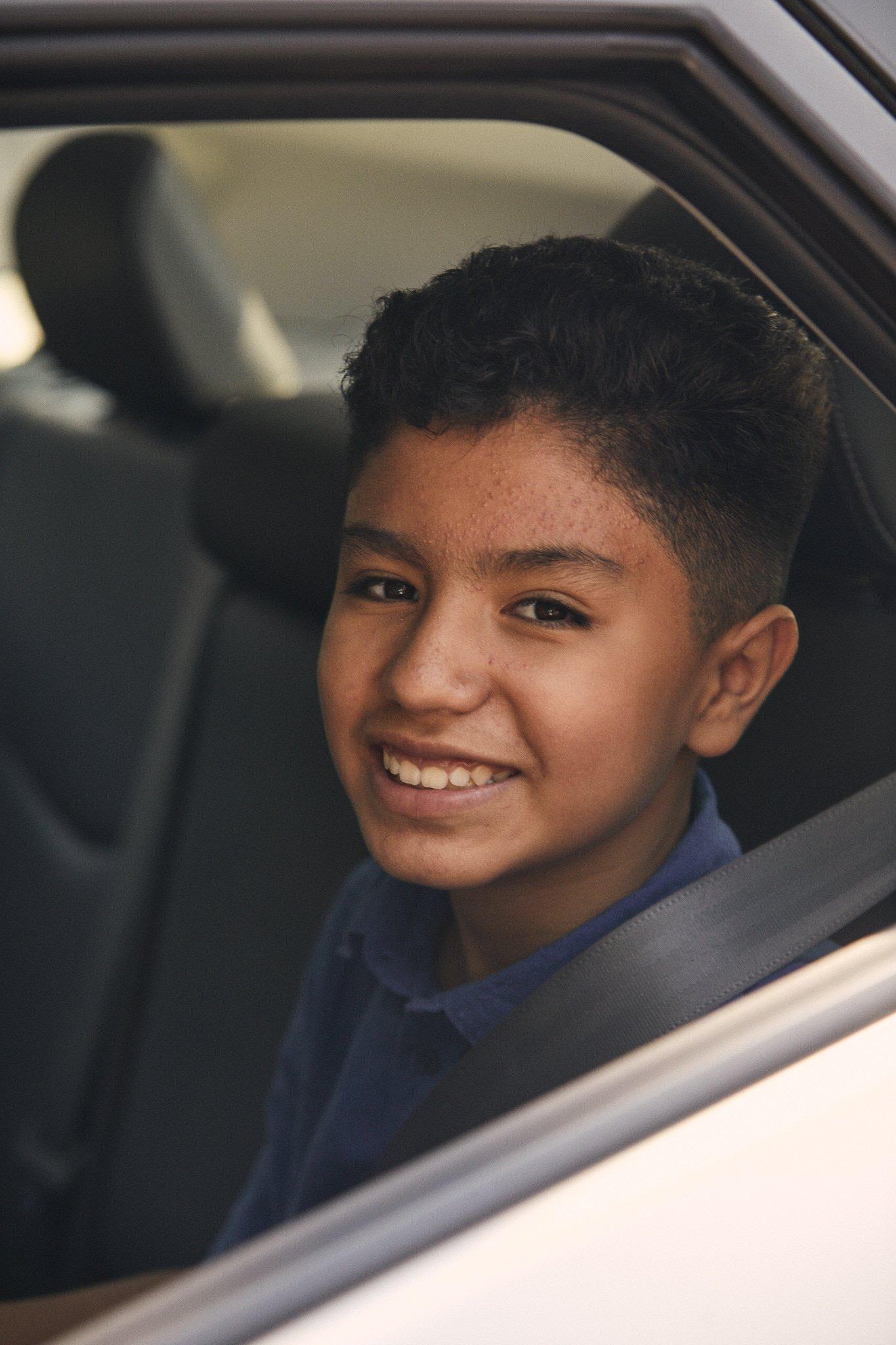 Smiling teenager in navy blue shirt sitting in car, viewed through car window with natural lighting