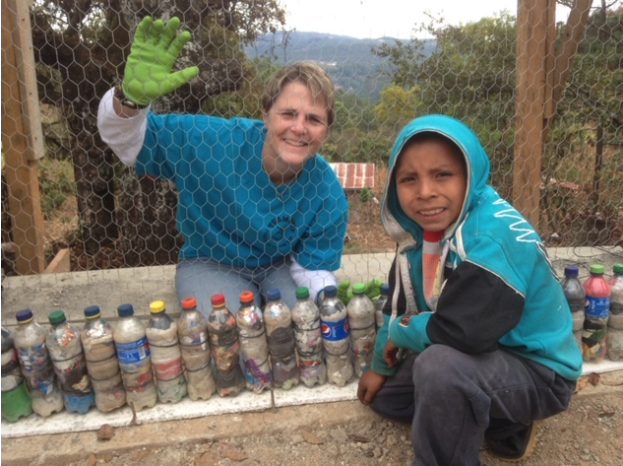 Two people posing next to a row of recycled plastic bottles used in a construction project, wearing blue hooded jackets and work gloves
