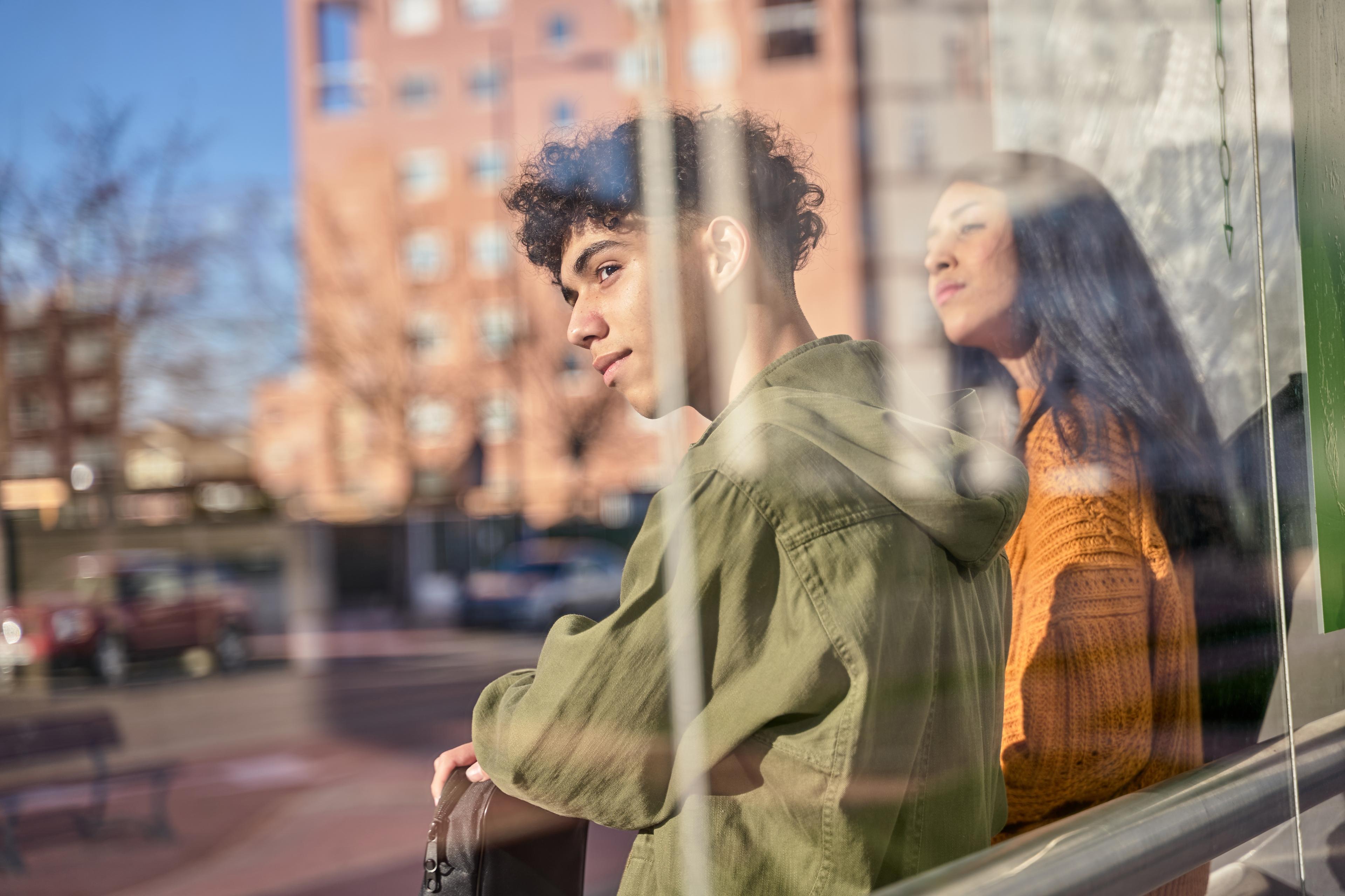 Two young people gazing through window in urban setting, one wearing green jacket, other in orange sweater with city backdrop