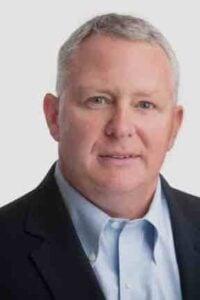 Professional headshot of a middle-aged man wearing a dark suit jacket and light blue dress shirt against a neutral background