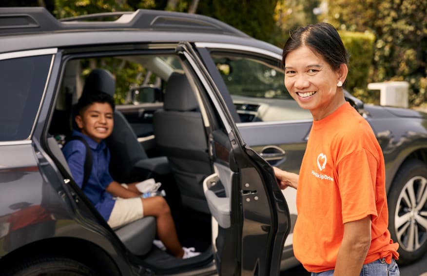 A driver wearing an orange company t-shirt opens the car door for a young passenger in the back seat of a modern SUV