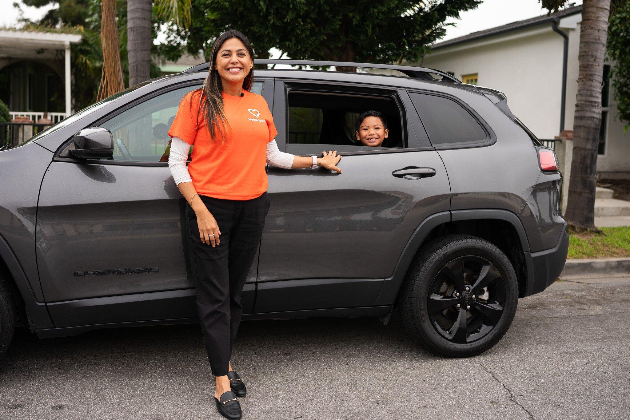 Driver in orange company t-shirt standing by grey Jeep Cherokee with young passenger visible in back window