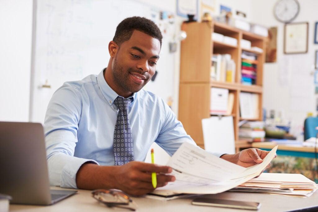 Professional in business attire reviewing paperwork at desk with laptop and office materials in organized workspace