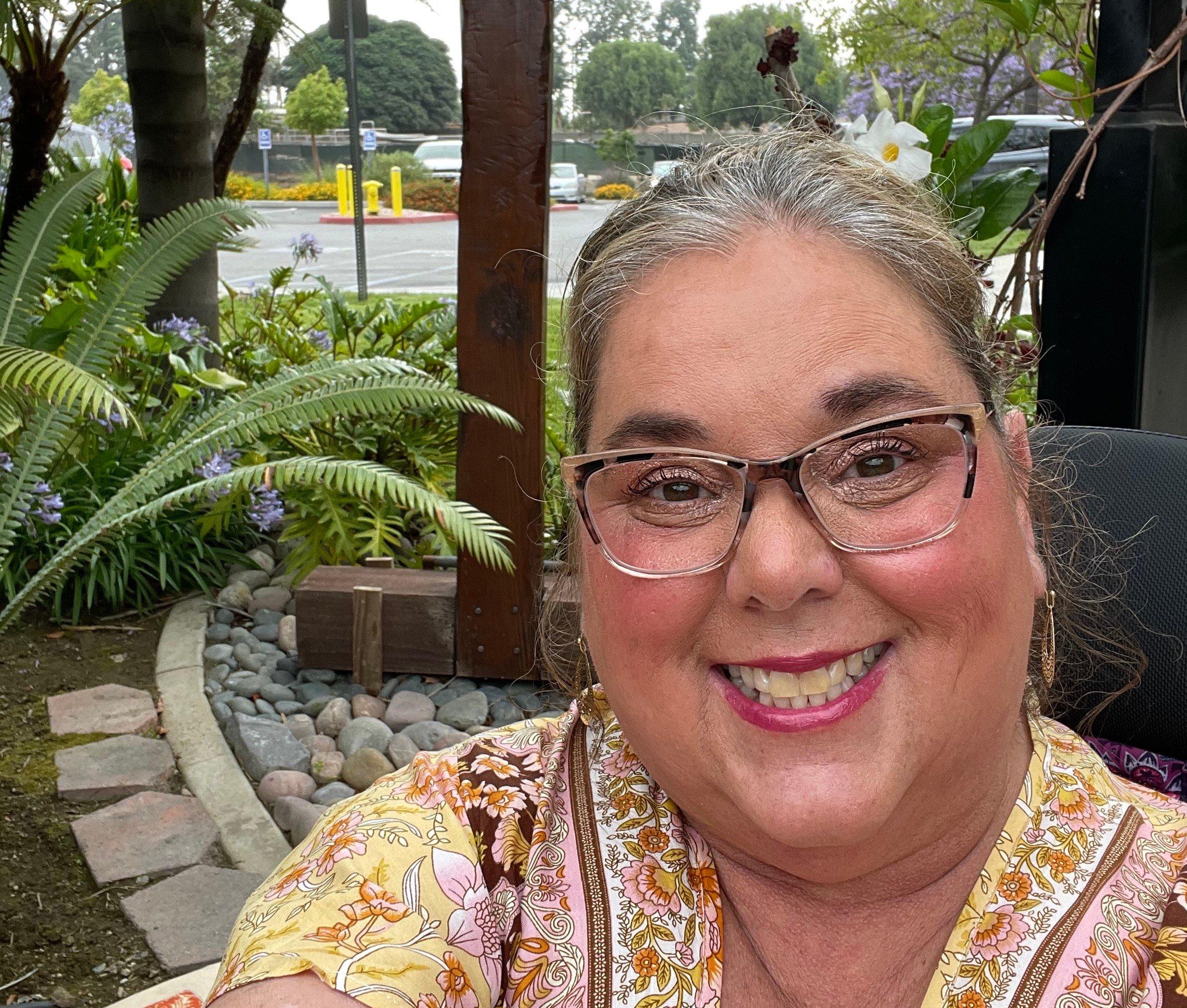 Woman with glasses and floral dress smiling warmly at camera in outdoor garden setting with ferns and stone landscaping