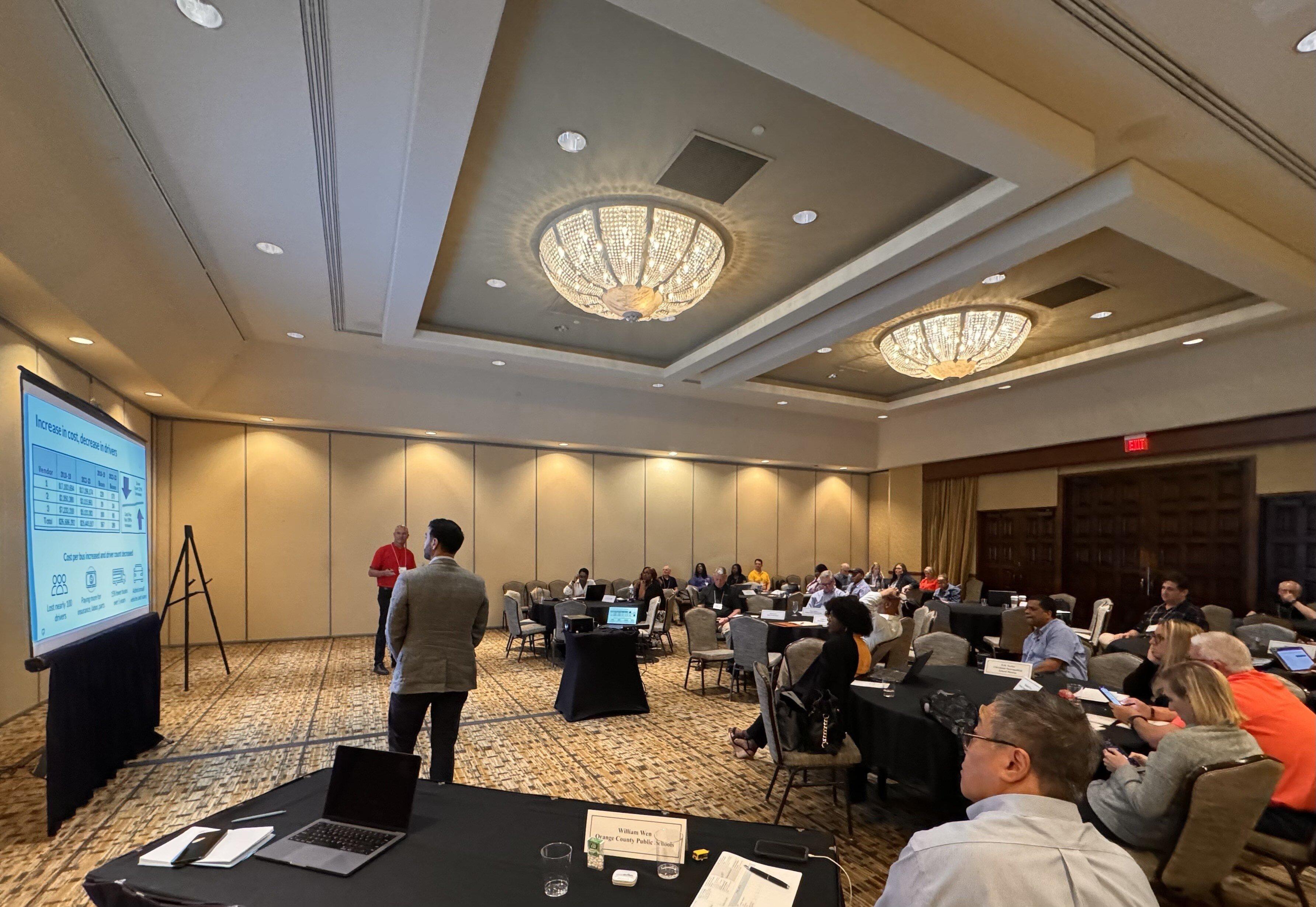 Business conference room with presentation screen, speakers at front, and attendees seated at tables with laptops